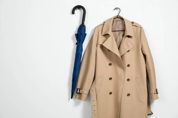 Hanger with beige trench coat and blue umbrella on white wall, space for text