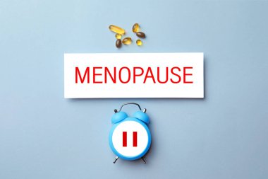 Menopoz kelimesi, duraklat sembollü alarm saati ve açık mavi arkaplanda haplar, üst görünüm