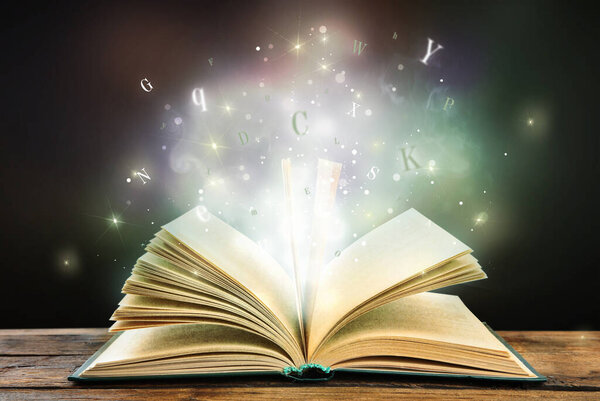Open book with magic light and glowing letters flying out of it on wooden table against black background
