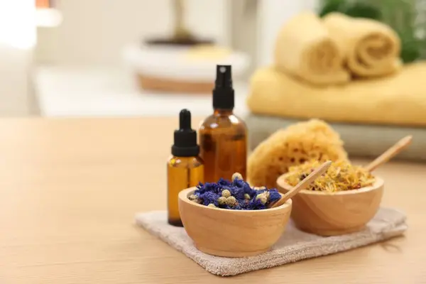 Bottles of essential oils, bowls with dry flowers and natural sponge on light wooden table, space for text. Spa therapy