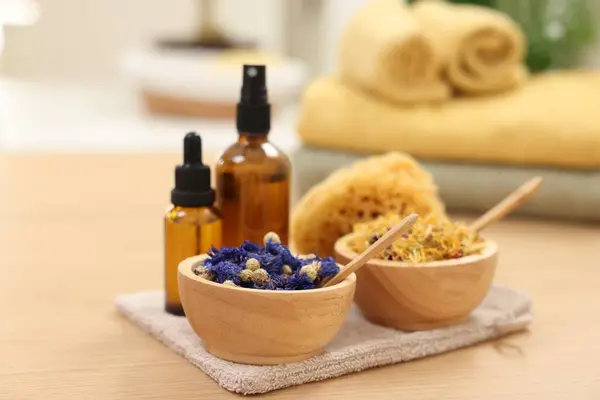 Bottles of essential oils, bowls with dry flowers and natural sponge on light wooden table, closeup. Spa therapy