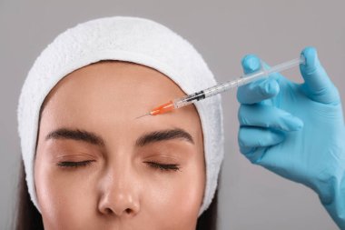 Doktor açık gri arka planda genç bir kadına yüz enjeksiyonu yapıyor, yakın plan. Estetik ameliyat