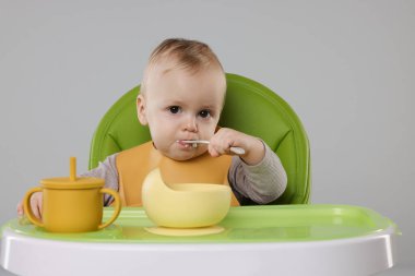 Gri arka planda, yüksek sandalyede sağlıklı yiyecekler yiyen şirin bir bebek.