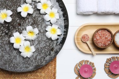 Çiçekli bir kase su ve beyaz ahşap masada farklı spa malzemeleri.