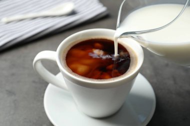 Gri masadaki sıcak kahve fincanına süt dökerek.