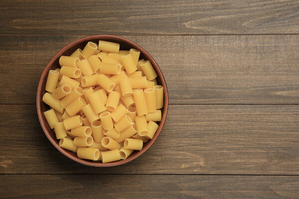 Raw rigatoni pasta in bowl on wooden table, top view. Space for text