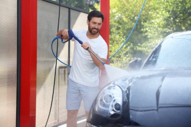 Dışarıdaki oto yıkamada yüksek basınçlı su jetiyle araba yıkayan mutlu bir adam.