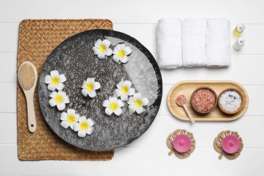Çiçekli bir kase su ve beyaz ahşap masada farklı spa malzemeleri.
