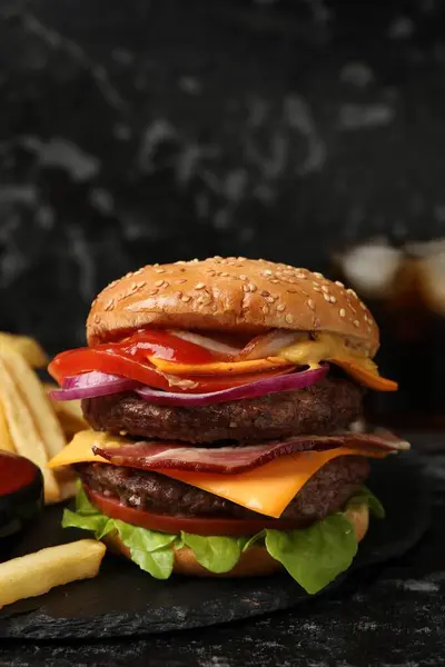 Delicious burger with meat cutlets, bacon, cheese, vegetables and lettuce on black table