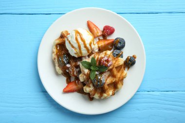 Açık mavi ahşap masada dondurma, böğürtlen ve karamel soslu leziz Belçika waffleları.