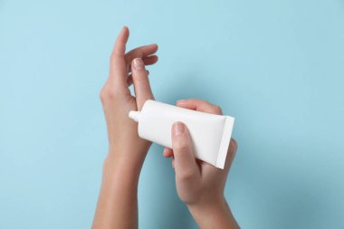 Kadın tüpten eline kozmetik krem sürüyor açık mavi arka planda, üst görünümde.