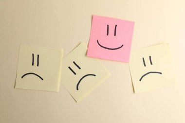 Seçim konsepti. Sarı kağıtlar arasında mutlu ifadeler içeren pembe yapışkan bir not bej arka planda üzgün emojiler, üst görünüm