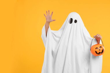 Beyaz hayalet kostümlü kadın sarı arka planda balkabağı kovası tutuyor, mesaj için yer var. Cadılar Bayramı