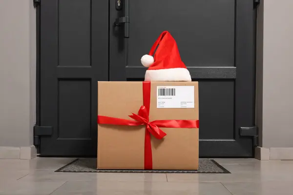 Christmas gift box and Santa hat near door indoors. Sending present by mail