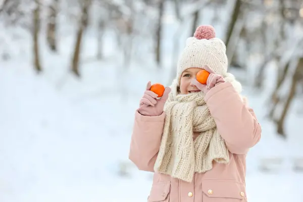 Cute little girl covering eye with tangerine in snowy park on winter day, space for text