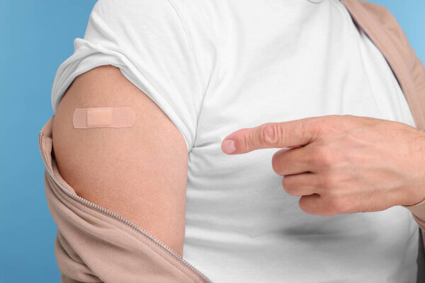 Man pointing at adhesive bandage after vaccination on light blue background, closeup