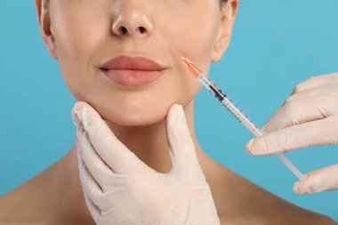 Doktor açık mavi arka planda genç bir kadına yüz enjeksiyonu yapıyor, yakın plan. Estetik ameliyat