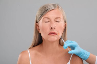 Doktor, açık gri arka planda yaşlı kadına yüz enjeksiyonu yapıyor. Yakın plan. Estetik ameliyat