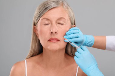 Doktor, açık gri arka planda yaşlı kadına yüz enjeksiyonu yapıyor. Yakın plan. Estetik ameliyat