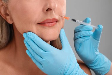 Doktor, açık gri arka planda yaşlı kadına yüz enjeksiyonu yapıyor. Yakın plan. Estetik ameliyat