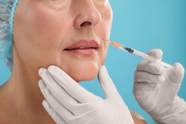 Doktor, açık mavi arka planda yaşlı kadına yüz enjeksiyonu yapıyor. Yakın plan. Estetik ameliyat