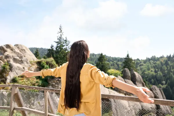 Feeling freedom. Woman with wide open arms near wooden railing in mountains, back view