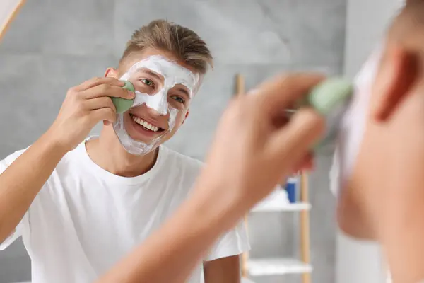 Happy young man washing off face mask with sponge near mirror in bathroom