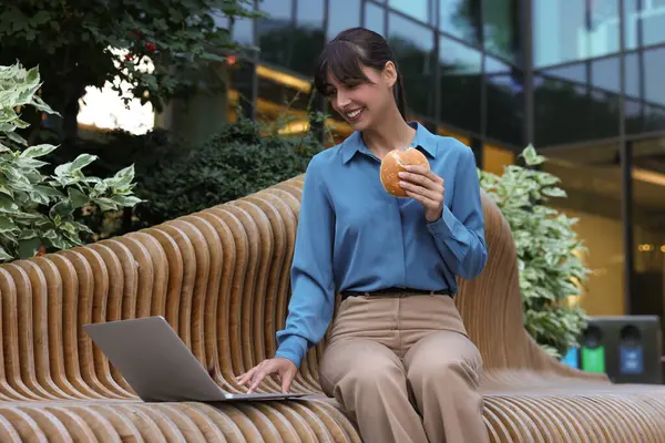 Lunch time. Happy businesswoman with hamburger using laptop on bench outdoors