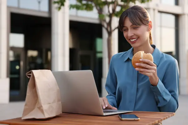 Happy businesswoman with hamburger using laptop during lunch at wooden table outdoors
