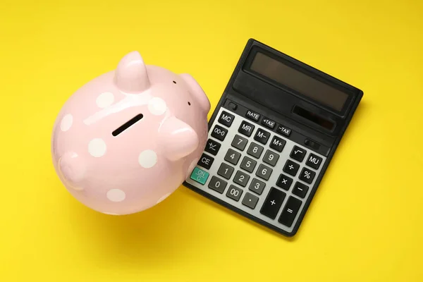 Calculator and piggy bank on yellow background, top view
