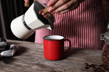 Moka pot 'tan gelen aromatik kahveyi, kapalı oturulan ahşap masada fincana döken kadın.