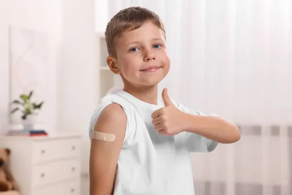 Boy with sticking plaster on arm after vaccination showing thumbs up indoors