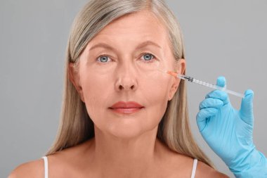 Doktor, açık gri arka planda yaşlı kadına yüz enjeksiyonu yapıyor. Yakın plan. Estetik ameliyat