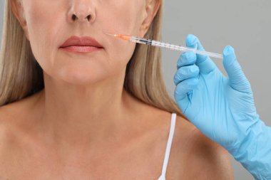 Doktor, açık gri arka planda yaşlı kadına yüz enjeksiyonu yapıyor. Yakın plan. Estetik ameliyat