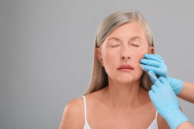 Doktor açık gri arka planda yaşlı kadına yüz enjeksiyonu yapıyor, yakın plan ve mesaj için yer. Estetik ameliyat