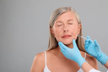 Doktor açık gri arka planda yaşlı kadına yüz enjeksiyonu yapıyor, yakın plan ve mesaj için yer. Estetik ameliyat