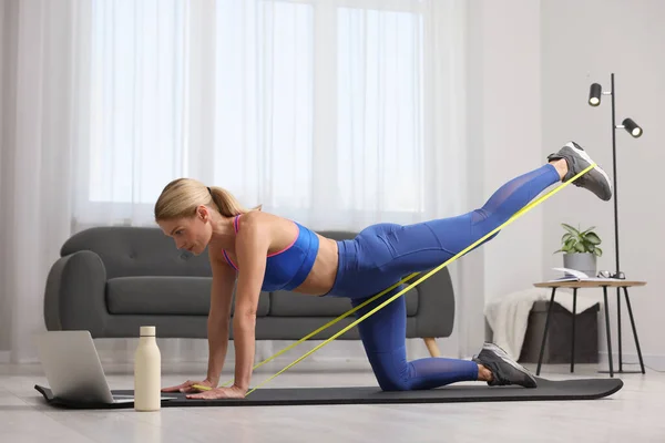 Fit woman doing exercise with fitness elastic band near laptop on mat at home