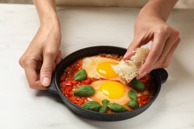 Beyaz masada Shakshuka 'nın içine bir parça ekmek daldıran kadın, yakın plan.