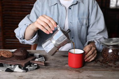 Moka pot 'tan gelen aromatik kahveyi, kapalı oturulan ahşap masada fincana döken kadın.