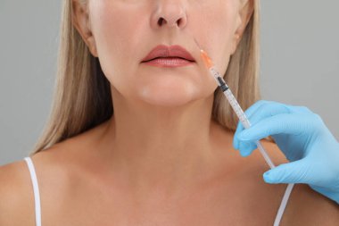 Doktor, açık gri arka planda yaşlı kadına yüz enjeksiyonu yapıyor. Yakın plan. Estetik ameliyat