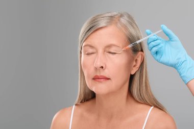 Doktor açık gri arka planda yaşlı kadına yüz enjeksiyonu yapıyor, yakın plan ve mesaj için yer. Estetik ameliyat