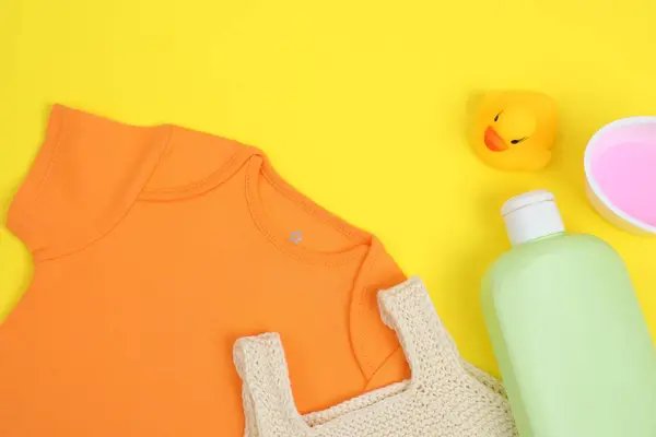 Laundry detergents, baby clothes and rubber duck on yellow background, flat lay