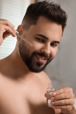 Yakışıklı bir adam banyoda yüzüne kozmetik serumu sürüyor.