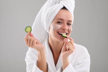 Yüzünde kozmetik ürünler ve gri arka planda salatalık dilimleri olan bir kadın. Spa tedavileri