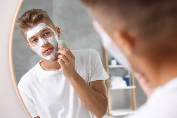 Young man washing off face mask with sponge near mirror in bathroom