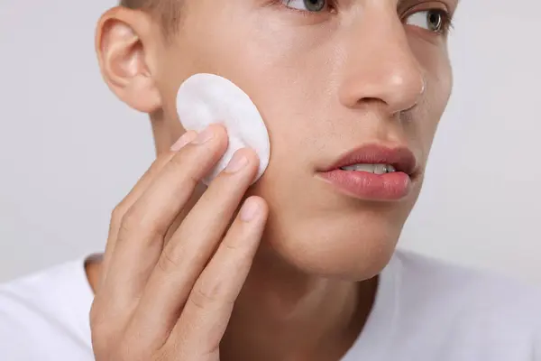 Man cleaning face with cotton pad on light background, closeup