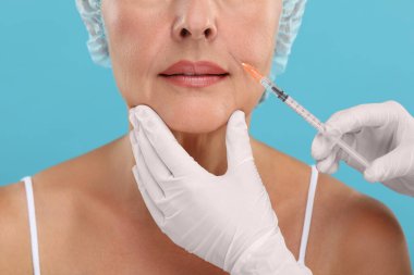 Doktor, açık mavi arka planda yaşlı kadına yüz enjeksiyonu yapıyor. Yakın plan. Estetik ameliyat