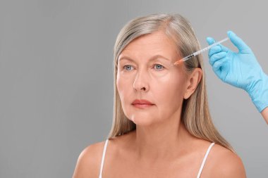 Doktor açık gri arka planda yaşlı kadına yüz enjeksiyonu yapıyor, yakın plan ve mesaj için yer. Estetik ameliyat