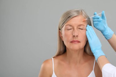Doktor açık gri arka planda yaşlı kadına yüz enjeksiyonu yapıyor, yakın plan ve mesaj için yer. Estetik ameliyat