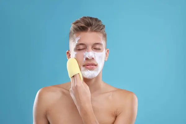 Young man washing off face mask with sponge on light blue background